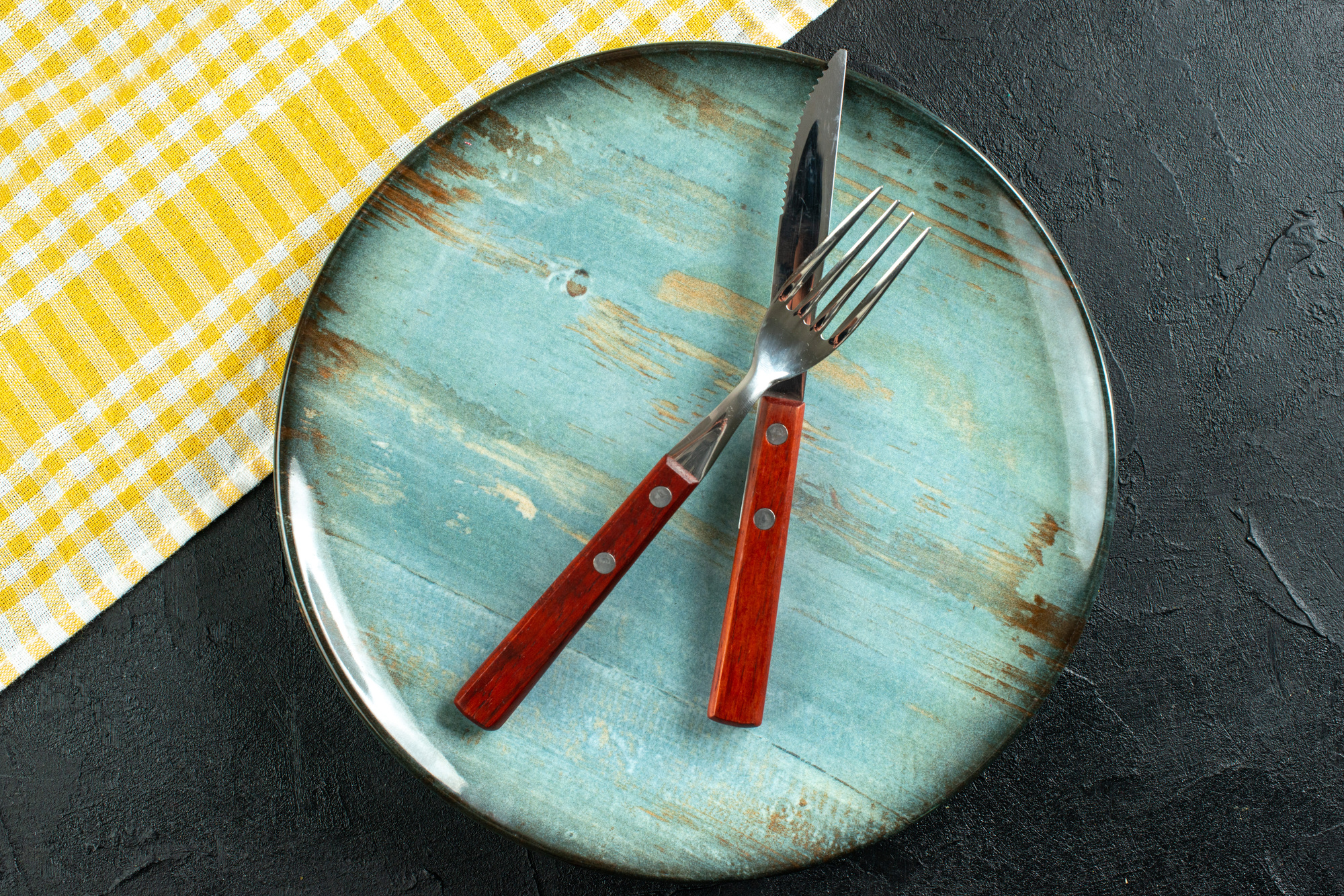 Horizontal view of meal cutlery in cross on a blue plate and yellow stripped towel on dark